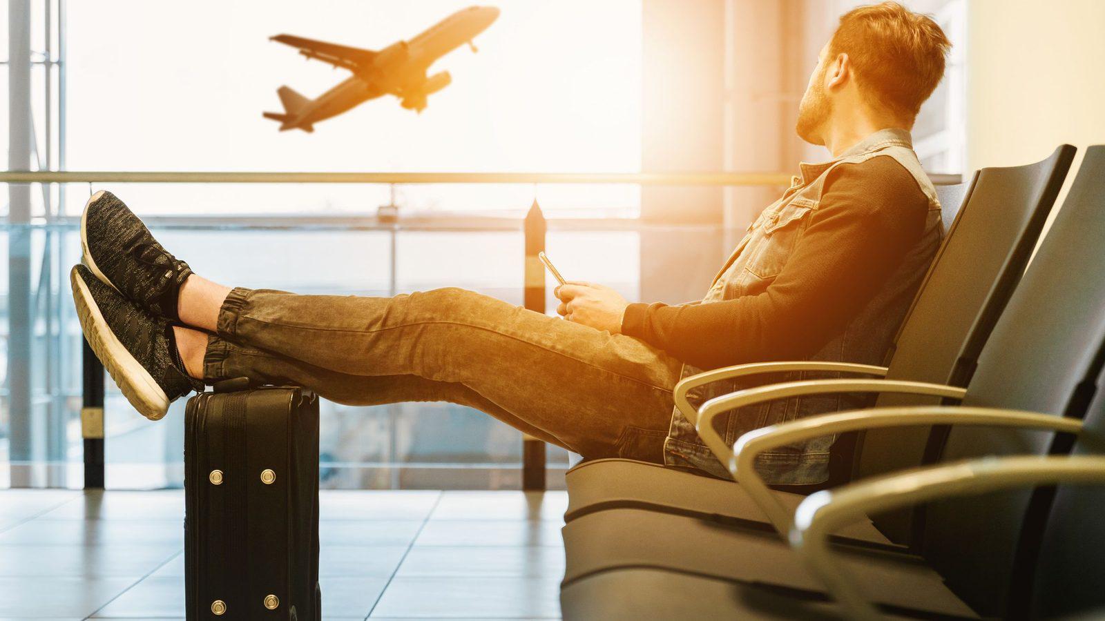 Un viajero espera su vuelo en un aeropuerto con el teléfono en la mano, mientras un avión despega al fondo. Foto de JESHOOTS.com en Pexels.